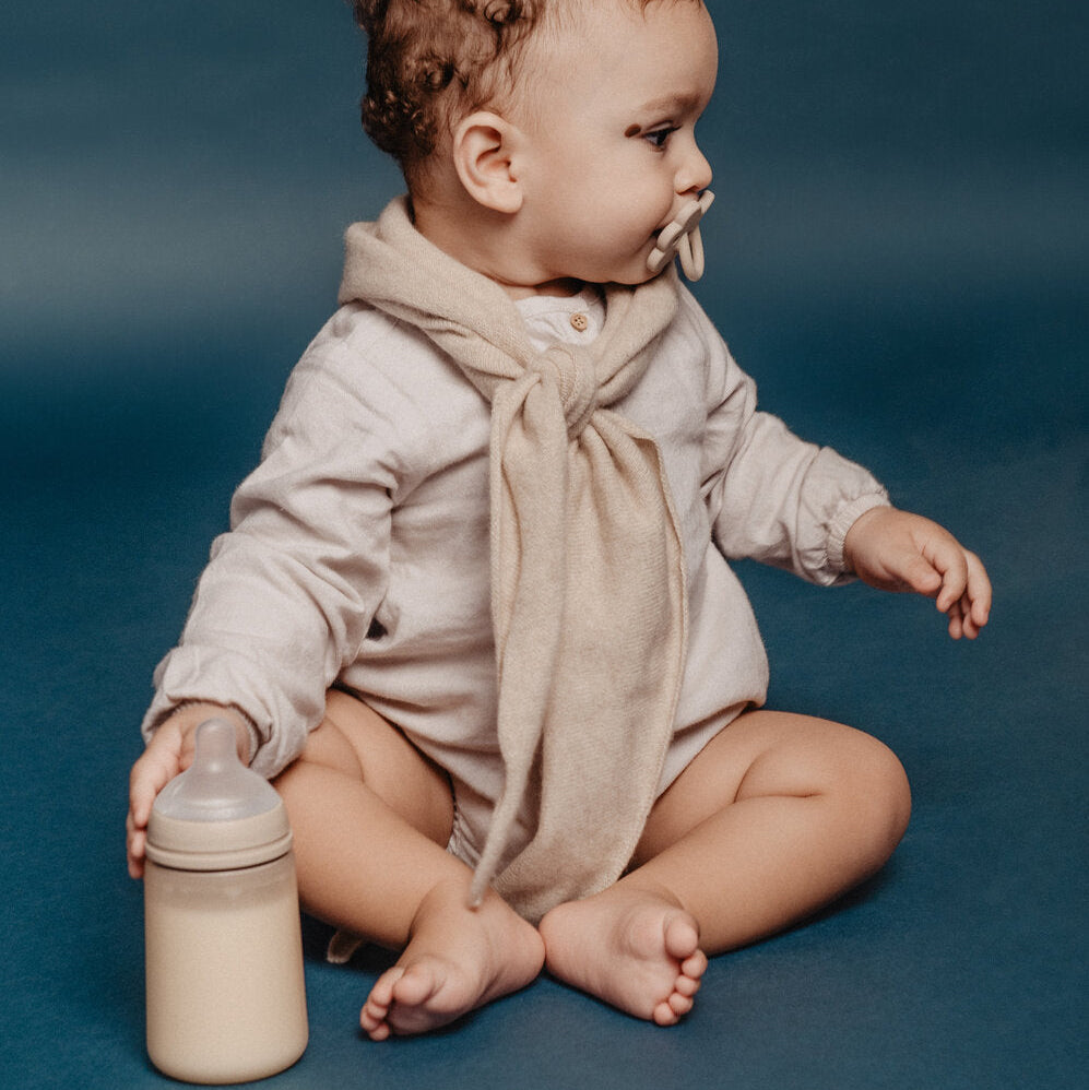 Baby sitting on a blue surface with a beige suavinex bottle and beige scarf