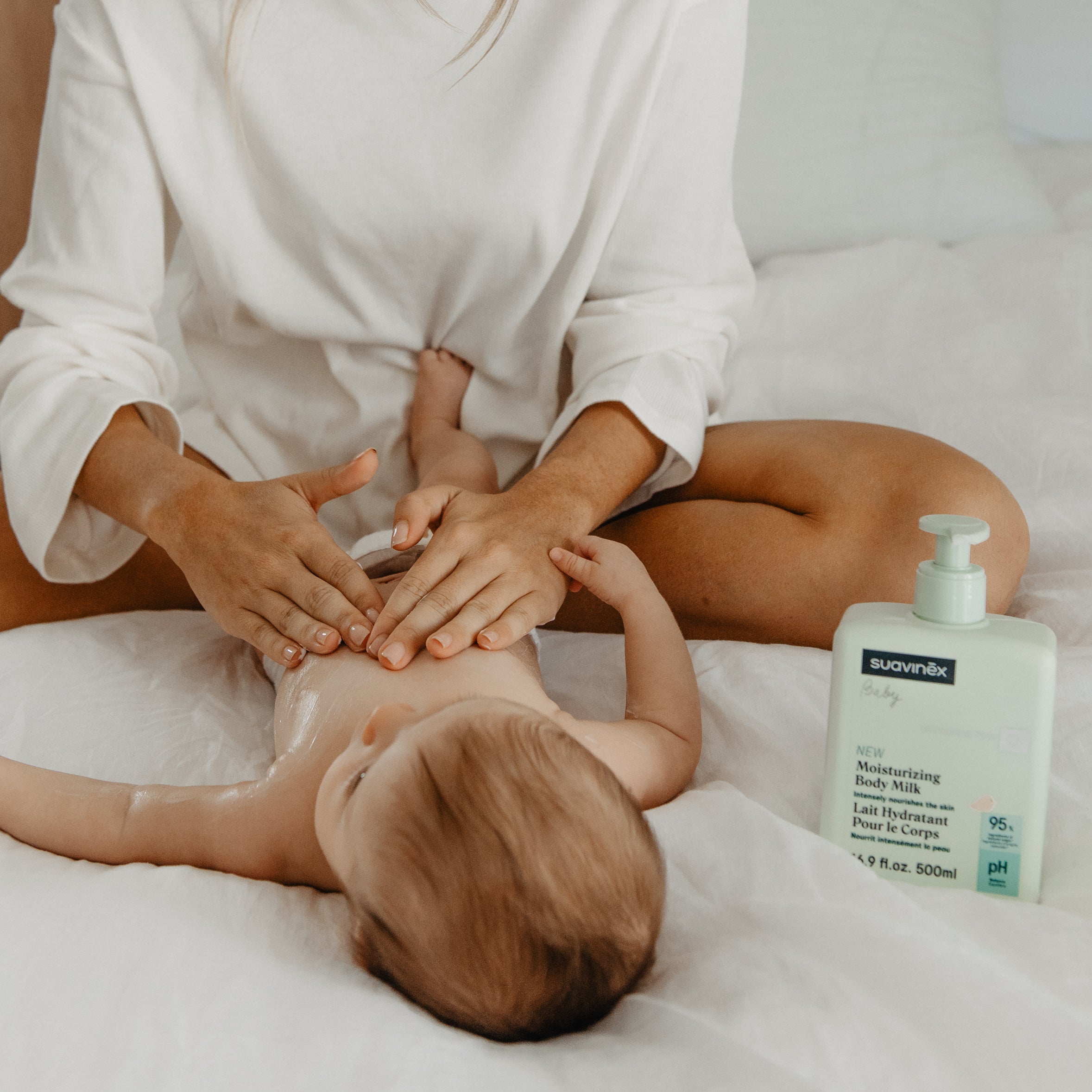 Mom applying suavinex moisturizing body milk to a baby's skin on a bed